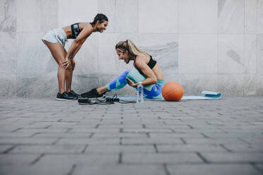 Seitenansicht einer Frau, die Sit-Ups macht, während ihre Freundin sich mit den Händen auf den Knien entspannt. Zwei Fitness-Frauen in fröhlicher Stimmung beim Training im Freien. - JLPSF20995