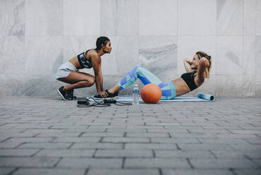 Fitness woman doing sit ups on exercise mat outdoors while her friend holds her legs. Women in fitness wear training outdoors. - JLPSF20996