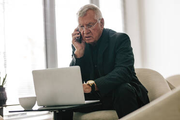 Älterer Geschäftsmann, der an einem Laptop arbeitet und mit einem Mobiltelefon in einer Büro-Lobby spricht. Ältere männliche Führungskraft, die in einer Büro-Lounge arbeitet. - JLPSF20998