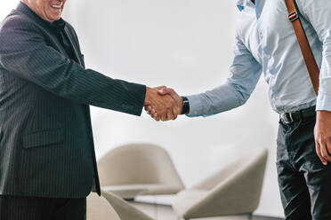 Zwei Geschäftsleute schütteln sich im Büro die Hände. Ein älterer Geschäftsmann begrüßt eine junge Führungskraft in der Büro-Lobby und heißt sie willkommen. - JLPSF20999
