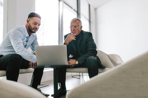 Aufnahme von zwei Geschäftsleuten, die in der Lobby eines modernen Büros einen Laptop benutzen. Ein junger Manager zeigt einem älteren Mann, der im Foyer sitzt, sein Geschäftsangebot. - JLPSF21000