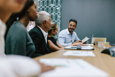 Positive young man with team of multi-ethnic people discussing productive strategy in boardroom. Executive reads a report for the business team in meeting room. - JLPSF21019