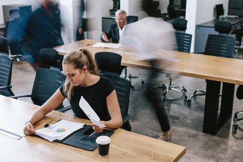 Geschäftsfrau sitzt am Tisch und prüft statistische Berichte. Weibliche Führungskraft arbeitet im Büro mit Menschen, die im Hintergrund vorbeigehen. - JLPSF21024