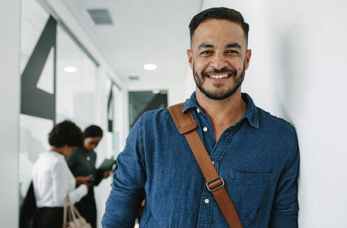 Portrait of young man in casuals standing at office corridor with colleagues in background. Male executive leaning to a wall in office hallway. - JLPSF21034