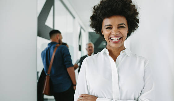 Portrait of happy young african businesswoman looking at camera and smiling with her team discussing in background. Cheerful woman in office hallway with coworkers at the back. - JLPSF21035