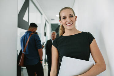 Portrait of happy young woman holding a laptop and smiling with her team discussing in background at office hallway. Happy businesswoman in office corridor with coworkers at the back. - JLPSF21037
