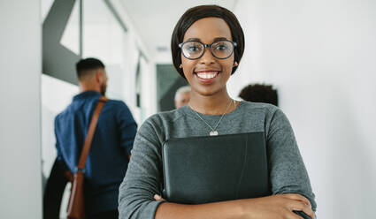 Porträt einer fröhlichen afrikanischen Geschäftsfrau mit Brille, die in einem Büro mit einem Team im Hintergrund steht. Lächelnde weibliche Führungskraft mit einem Ordner im Büroflur mit Mitarbeitern, die sich im Hintergrund unterhalten. - JLPSF21039