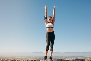 Lächelnde Frau in Fitnesskleidung, die auf einem Hügel mit klarem, blauem Himmel im Hintergrund steht und Übungen macht. - JLPSF21053