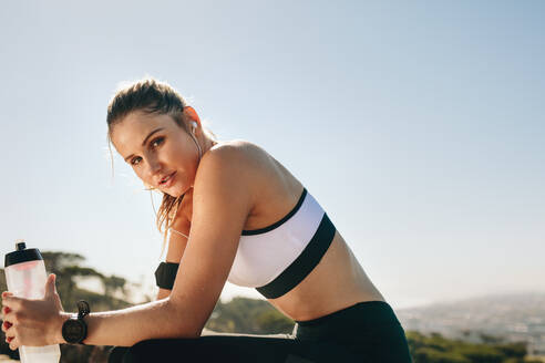 Seitenansicht einer Fitness-Frau, die im Freien sitzt und eine Wasserflasche hält. Frau in Fitnesskleidung, die sich nach dem Training entspannt, Wasser trinkt und Musik über Kopfhörer hört. - JLPSF21054