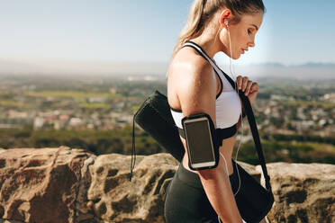 Seitenansicht einer Fitness-Frau, die nach dem Training mit ihrer Fitnesstasche auf einem Hügel läuft. Frau in Fitnesskleidung mit einem Mobiltelefon am Arm hört nach dem Training Musik. - JLPSF21055
