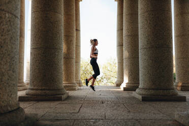 Frau in Fitnesskleidung beim Seilspringen in der Nähe von Steinsäulen. Seitenansicht einer Sportlerin beim Training mit dem Springseil. - JLPSF21061