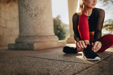 Woman in fitness wear sitting on ground and tying her shoelace. Fitness woman tightening her shoes during workout. - JLPSF21078