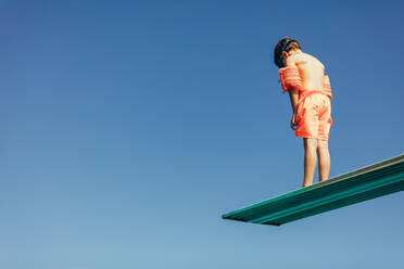 Low angle shot of boy with sleeves floats on diving board preparing for dive in the pool. boy standing on diving spring board against sky. - JLPSF21085