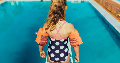 Rear view of girl standing on spring board learning to dive at swimming class. Girl learning spring board diving at outdoor pool. - JLPSF21086