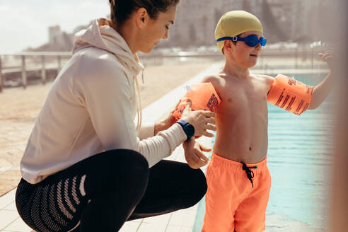 Weiblicher Trainer, der einem Jungen beim Anziehen der Schwimmflügel hilft; Frau, die dem Jungen die Schwimmflügel für den Schwimmunterricht am Pool anzieht. - JLPSF21090