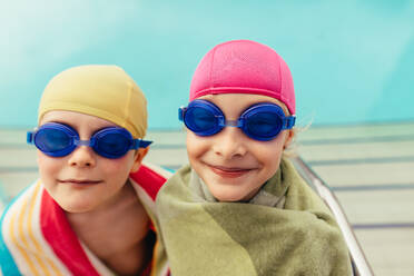 Junge und Mädchen mit Schwimmbrille und Kappe, eingewickelt in ein Handtuch, nach dem Schwimmtraining. Zwei Kinder, eingewickelt in ein Handtuch, am Pool. - JLPSF21094