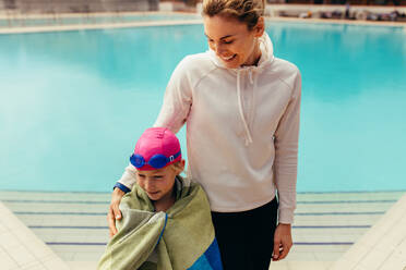 Mädchen, eingewickelt in ein Handtuch, steht mit einer Frau am Schwimmbecken. Mädchenkind mit Trainer am Schwimmbecken. - JLPSF21096