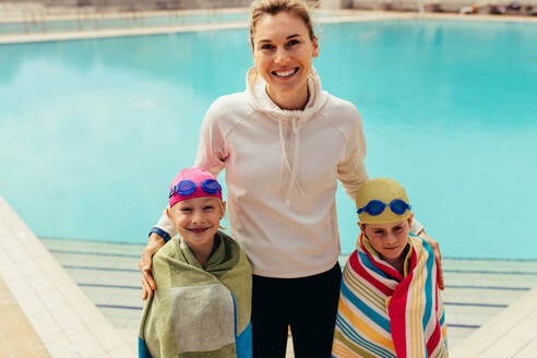 In Handtücher eingewickelte Kinder stehen mit einer Trainerin am Schwimmbecken. Mädchen und Junge mit Trainer am Schwimmbecken. - JLPSF21097