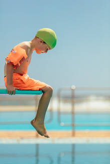 Junge auf einem Sprungturm sitzend und in das Schwimmbecken blickend. Junge in Badekleidung auf dem Sprungbrett sitzend. - JLPSF21107
