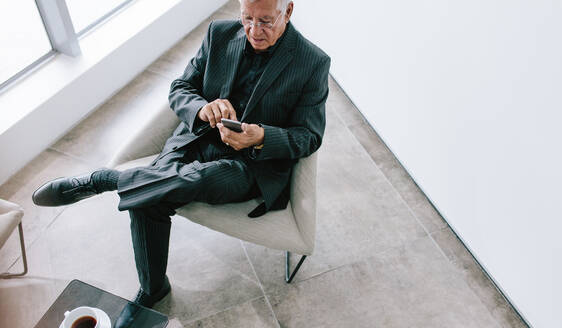 Hoher Winkel Blick auf ältere Geschäftsmann SMS mit Handy. Ältere Unternehmen professionelle Lesen von E-Mails auf seinem Smartphone während der Pause. Älterer Mann in Business-Anzug sitzt in Büro-Lobby mit Handy in Büro-Lobby. - JLPSF21112