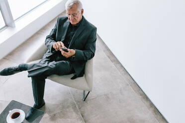 Blick auf einen alten Geschäftsmann, der in der Büro-Lobby sein Mobiltelefon benutzt. Ein reifer Geschäftsmann liest in der Pause eine Textnachricht auf seinem Smartphone. - JLPSF21113