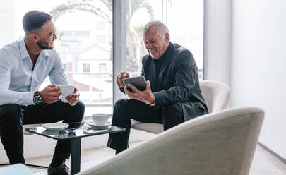 Lächelnder älterer Geschäftsmann mit digitalem Tablet, der mit einem jungen männlichen Kollegen in der Büro-Lobby diskutiert. Zwei Geschäftsleute trinken Kaffee und unterhalten sich zwanglos im Büro-Foyer. - JLPSF21115