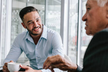 Young businessman talking with senior man over a cup of coffee. Business people having casual discussion during coffee break. - JLPSF21117