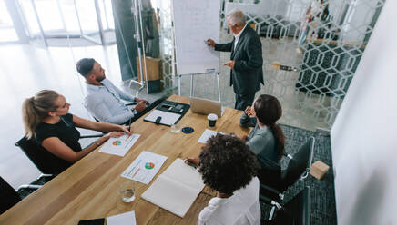 Mature businessman presenting his ideas on whiteboard to colleagues. Senior manager giving presentation to his team sitting in conference room at office - JLPSF21122