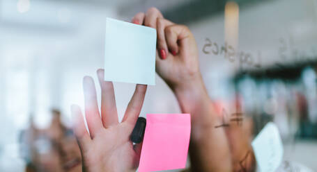 Hands of woman pasting sticky notes on glass wall in office. Business people discussing new ideas with post it notes on transparent glass wall. - JLPSF21133