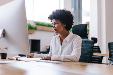 Junge afrikanische Frau sitzt an ihrem Schreibtisch, schaut auf den Computerbildschirm und macht sich Notizen in einem Buch. Eine Unternehmerin arbeitet in ihrem Büro. - JLPSF21136