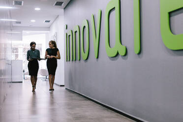 Full length of two business women walking through office hallway. Female coworkers walking in office corridor. - JLPSF21144