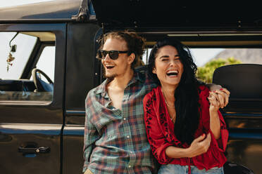 Fröhliches junges Paar, das auf einem Roadtrip eine Pause einlegt. Junger Mann und Frau stehen bei ihrem Auto und lachen. - JLPSF21154