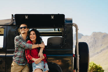 Couple standing beside their car on a highway. Man embracing his woman while standing outdoors and looking away smiling. - JLPSF21156