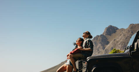 Junger Mann sitzt mit seiner Freundin auf der Motorhaube eines Autos, das im Freien steht, mit einem Berg im Hintergrund. Romantisches junges Paar, das bei seinem Auto steht und die Natur genießt. - JLPSF21158