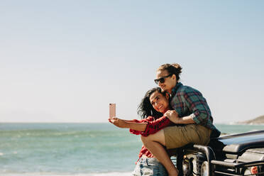 Verliebtes junges Paar macht ein Selfie während eines Roadtrips. Mann und Frau stehen am Strand neben ihrem Auto und machen ein Selfie. - JLPSF21162