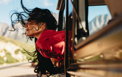 Beautiful woman sitting on car with her face out of the window with her hair flying in air. Female enjoying the car ride on a road trip. - JLPSF21182