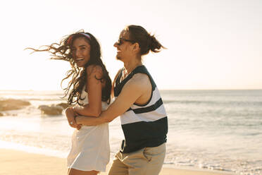Glückliches junges Paar, das sich im Sommerurlaub am Strand amüsiert. Junger Mann hält seine Freundin von hinten und lächelt. - JLPSF21188