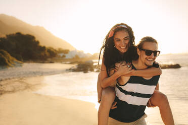 Junger Mann und Frau im Urlaub an einem schönen Sommertag. Glücklicher Mann, der eine Frau auf seinem Rücken trägt und am Strand spazieren geht. - JLPSF21193