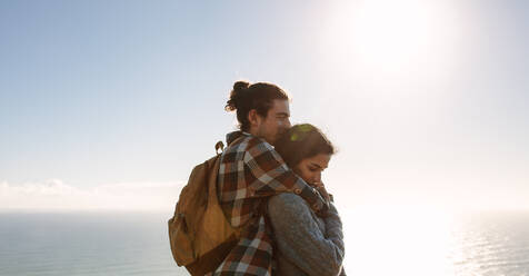 Junges verliebtes Paar an einem sonnigen Tag im Freien mit dem Meer im Hintergrund. Mann mit Rucksack umarmt Frau. - JLPSF21205