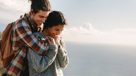 Romantischer junger Mann, der seine Freundin von hinten umarmt und lächelt. Verliebtes Paar, das im Freien steht, mit dem Meer im Hintergrund. - JLPSF21209