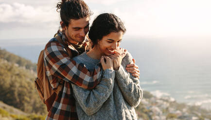 Porträt eines schönen jungen Paares im Urlaub: Mann umarmt seine Freundin von hinten, beide stehen auf einer Klippe. - JLPSF21210