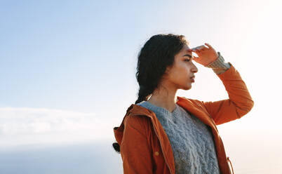 Portrait of young woman standing outdoors and looking away with hand on forehead. Female in warm clothing staring at a view. - JLPSF21216