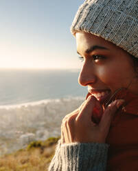 Close up of smiling african woman enjoying the beautiful sea view from mountain top. Female in warm jacket and knitted cap. - JLPSF21231