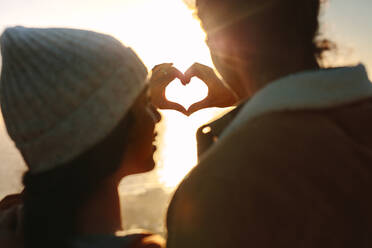 Rear view close up of man and woman making heart with hands against bright sunshine. Couple in love making heart with fingers. - JLPSF21240