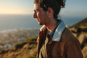 Porträt eines jungen Mannes mit Jacke, der auf einer Klippe steht und die Aussicht genießt; ein Mann im Urlaub an einem Wintertag. - JLPSF21242