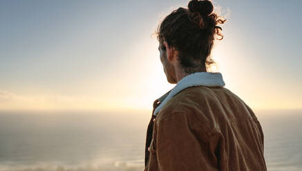 Rückansicht eines jungen Mannes mit Blick auf das Meer. Mann in warmer Jacke betrachtet die Aussicht von einem Berg aus. - JLPSF21243