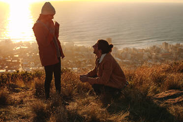 Junger Mann kniet auf dem Boden mit einer Frau, die auf einer Klippe steht. Verliebtes Paar in den Bergen mit Meereslandschaft im Hintergrund. - JLPSF21244