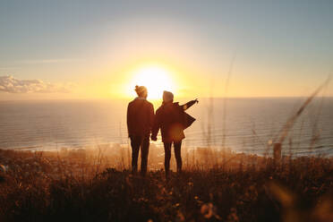 Silhouette eines jungen Mannes und einer Frau, die auf einem Berg stehen, wobei die Frau auf das Meer zeigt. Das Paar betrachtet die Meereslandschaft während des Sonnenuntergangs. - JLPSF21250