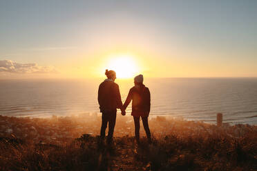Silhouette eines Mannes und einer Frau, die sich an den Händen halten und auf dem Berggipfel bei Sonnenuntergang stehen. Reisendes Paar auf einer Klippe bei Sonnenuntergang. - JLPSF21252