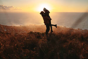 Silhouette eines jungen Mannes, der seine Freundin auf den Berggipfel trägt, mit einer Meereslandschaft im Hintergrund. Romantisches Paar, das sich auf einer Klippe bei Sonnenuntergang vergnügt. - JLPSF21255